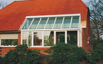 Klassischer Wintergarten mit schrägem Glasdach und weißen Rahmen, angebaut an ein Haus mit rotem Ziegeldach.