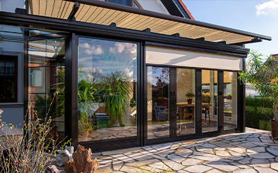 Wintergarten mit großen Glasflächen, außenliegendem Sonnenschutz und hellem Wohnraum mit Pflanzen.