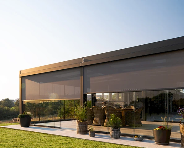 Großflächige Glasfassade mit textilen Screens als Sonnenschutz, Terrasse mit Gartenmöbeln im Abendlicht.