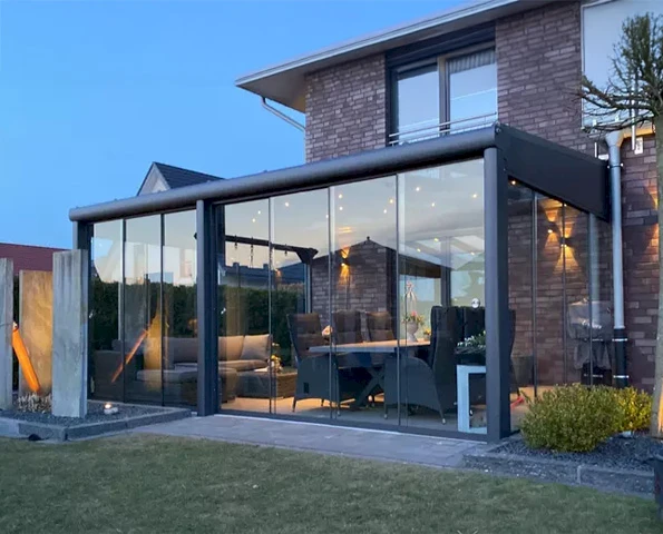 Sommergarten mit rahmenloser Verglasung und stilvoller Einrichtung, stimmungsvoll beleuchtet in der Dämmerung.