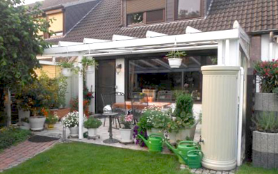 Weiße Terrassenüberdachung mit Glasdach und gemütlicher Sitzecke im Garten.