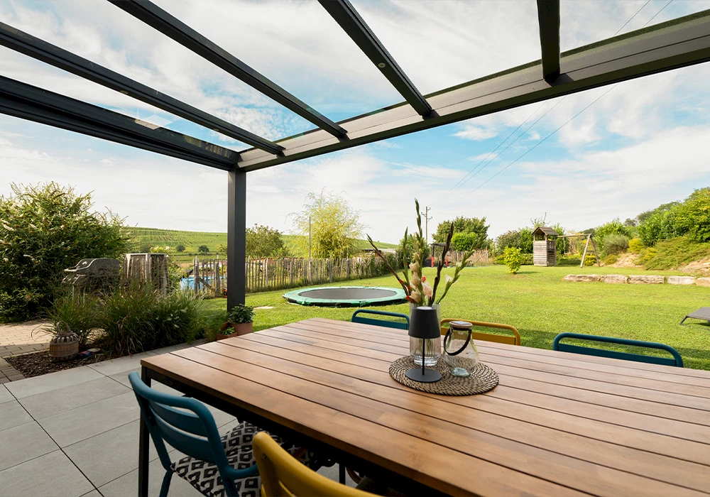Terrassenüberdachung aus Glas mit Beschattung und Blick auf Garten mit Spielgeräten und Trampolin