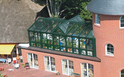 Zweigeschossiger Wintergarten mit grünem Aluminiumrahmen und Glasdach, angebaut an ein Backsteingebäude.