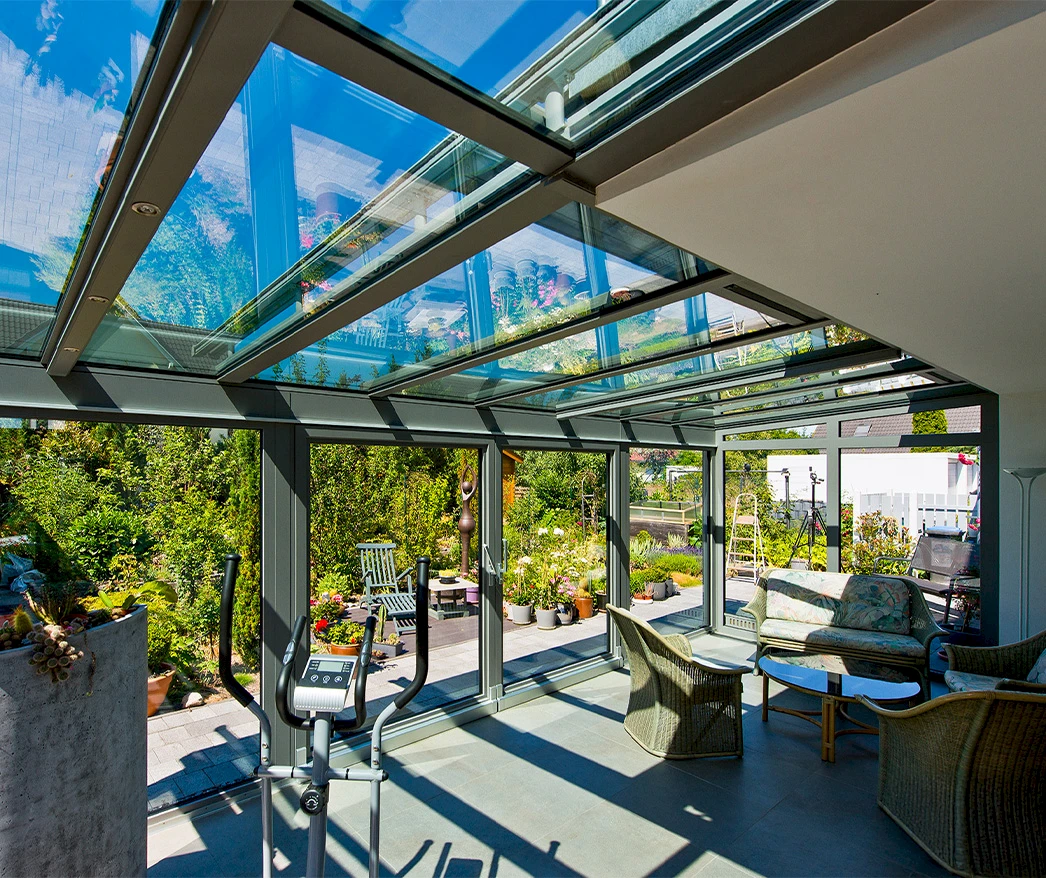 Heller Wintergarten mit Glasdach, Sitzmöbeln und Blick in den Garten, viel Tageslicht durch große Fenster.