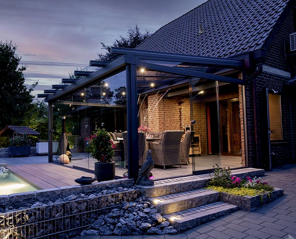 Sommergarten mit rahmenloser Verglasung und stilvoller Einrichtung, stimmungsvoll beleuchtet in der Dämmerung.