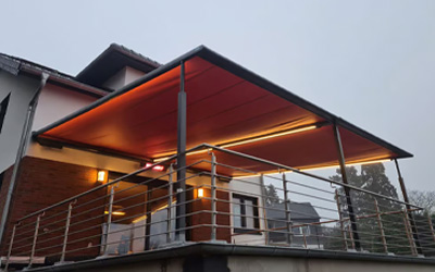 Terrassenüberdachung mit rotem Tuch und LED-Beleuchtung auf erhöhter Terrasse mit Edelstahlgeländer.