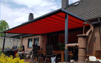 Terrassenüberdachung mit rotem Markisentuch über gemütlichem Sitzbereich an einem Haus mit Ziegeldach.