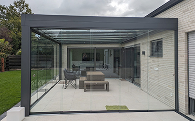 Sommergarten mit rahmenlosen Glasschiebewänden und Glasdach an einem Neubau mit heller Terrasse.