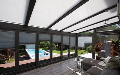 Wintergarten-Innenraum mit Sonnenschutz, bodentiefen Fenstern und Blick auf den Garten mit Pool.