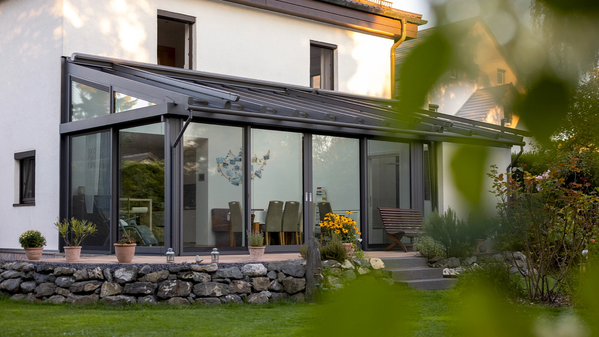 Angebauter Wintergarten mit Pultdach aus Aluminium und großflächiger Verglasung, mit Holzterrasse und gepflegtem Garten