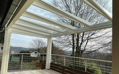 Weiße Terrassenüberdachung mit Glasdach auf einer erhöhten Terrasse mit Geländer und Ausblick ins Grüne.