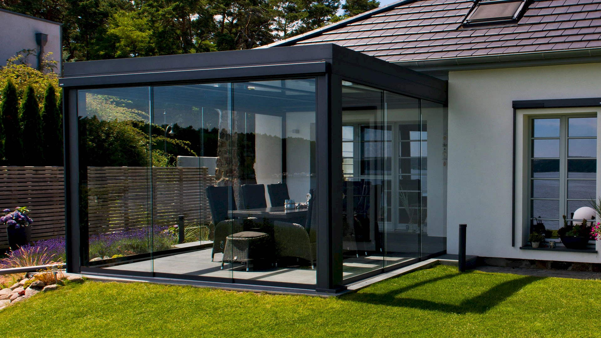 Moderner Sommergarten an einem Einfamilienhaus mit rahmenlosen Glasschiebetüren, Essbereich und kompakter Terrasse.